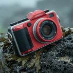 Appareil photo numérique rouge robuste de marque Luminova posé sur un rocher volcanique humide couvert d'algues en bord de mer.