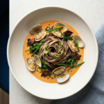 Dressage gastronomique d'un plat de pâtes au sarrasin avec coques, salicorne et morceaux d'andouille dans une sauce onctueuse.